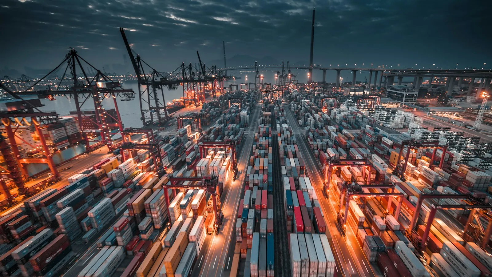 A container port at dusk, lit in orange, with cranes and stacked containers